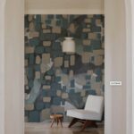 Una stanza spaziosa con un'ampia apertura ad arco che rivela una parete accenta con una carta da parati a motivi geometrici. La carta da parati presenta una combinazione di quadrati e rettangoli in varie tonalità di blu, grigio e beige, creando un motivo astratto e moderno. Al centro della stanza, sospesa al soffitto, c'è una lampada a sospensione bianca, semplice ma elegante. Una poltrona moderna in tessuto chiaro con braccioli in legno è posizionata sul lato destro della stanza, accanto a un piccolo tavolino rotondo in legno. Il pavimento è in pietra chiara, con un tappeto a motivi geometrici nei toni del grigio e del bianco che aggiunge un tocco di calore e texture. La stanza è inondata di luce naturale, creando un'atmosfera accogliente e rilassante.