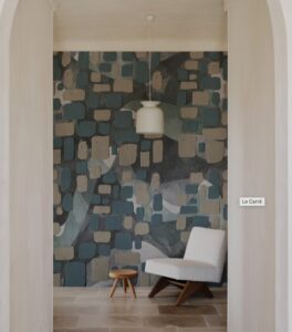 Una stanza spaziosa con un'ampia apertura ad arco che rivela una parete accenta con una carta da parati a motivi geometrici. La carta da parati presenta una combinazione di quadrati e rettangoli in varie tonalità di blu, grigio e beige, creando un motivo astratto e moderno. Al centro della stanza, sospesa al soffitto, c'è una lampada a sospensione bianca, semplice ma elegante. Una poltrona moderna in tessuto chiaro con braccioli in legno è posizionata sul lato destro della stanza, accanto a un piccolo tavolino rotondo in legno. Il pavimento è in pietra chiara, con un tappeto a motivi geometrici nei toni del grigio e del bianco che aggiunge un tocco di calore e texture. La stanza è inondata di luce naturale, creando un'atmosfera accogliente e rilassante.